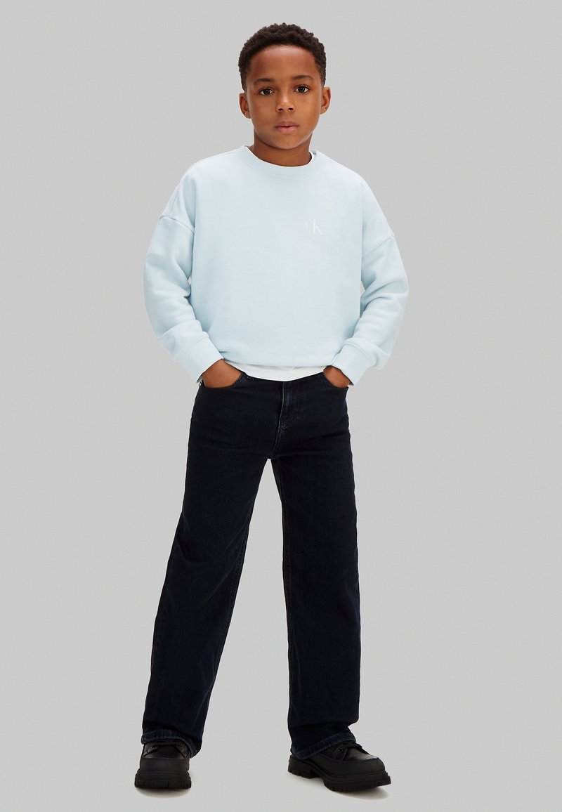 Garçon debout les mains dans les poches, portant un sweat-shirt bleu clair, un pantalon noir à jambes larges et des chaussures noires, sur un fond gris.