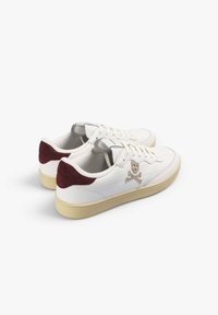 Zapatillas blancas con acentos de ante burdeos, que presentan detalles perforados, un logo de calavera y huesos cruzados, y una suela de goma crema.