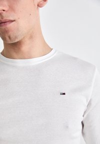 Vit långärmad t-shirt i bomull med rund halsringning. Har en liten broderad logotyp i rött, vitt och blått på bröstet.