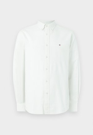 Bijela košulja dugih rukava od pamuka. Ima ovratnik s gumbima, prednje gumbe i mali logo na lijevoj strani prsa.