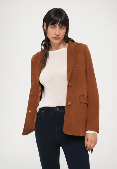 Blazer de camurça castanha com corte descontraído, design de um só peito, lapelas entalhadas e dois botões decorativos. Combinado com um sweater claro e jeans escuros.