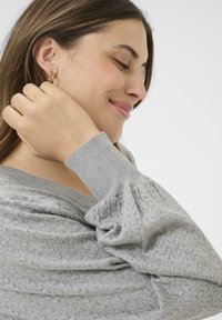 Šedý pletený svetr s texturovaným vzorem a žebrovanými manžetami, doplněný jednoduchými kruhovými náušnicemi. Modelka se dotýká výstřihu.