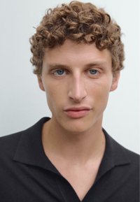 Detailní záběr osoby s kudrnatými světle hnědými vlasy a modrýma očima, která má na sobě černé polo tričko. Pozadí je čistě bílé.