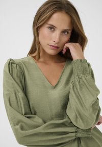 Blouse vert olive avec un décolleté en V, tissu texturé et manches bouffantes. Présente des détails smockés au niveau des poignets et de la taille pour un design ajouté.