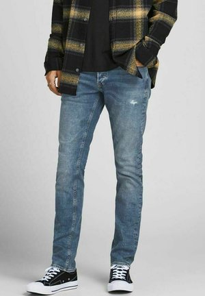 Vaqueros ajustados de color azul claro con un detalle desgastado en el frente, combinados con una camisa de cuadros negra y amarilla y zapatillas deportivas negras.