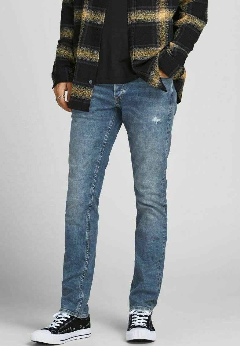 Světle modré slim-fit džíny s viditelným opotřebením na přední straně, spárované s černou a žlutou kostkovanou košili a černými teniskami.