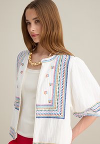 Chaqueta blanca bordada que presenta patrones geométricos coloridos a lo largo de los bordes y las mangas, con una tela texturizada y ligera.
