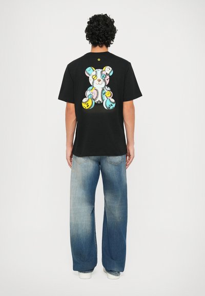 Juodas medvilnės marškinėlis su spalvingu meškino grafiku ant nugaros, derinamas su šviesiai mėlynomis plačiomis džinsinėmis kelnėmis.