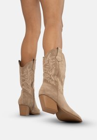 Botas de cowboy de ante beige con punteras afiladas, diseño floral bordado y tacones apilados, que destacan por su textura suave y costuras definidas.