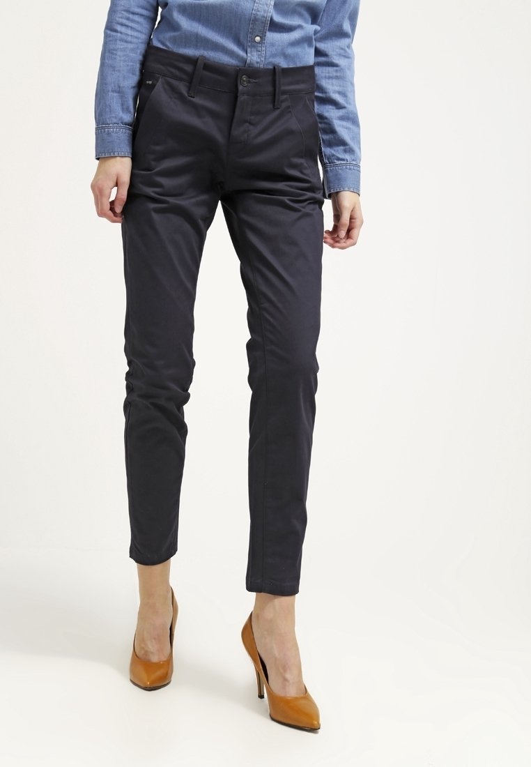 Femme portant un pantalon slim noir, une chemise bleue boutonnée et des chaussures à talons marron, debout devant un fond blanc.