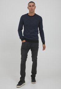 Navy blue knitted sweater featuring a diamond pattern, black ribbed collar and cuffs, worn with black slim-fit jeans and black shoes.