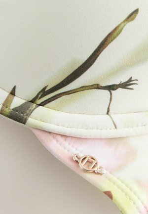 Audums ar ziedu rakstu tuvplānā un mazs, apaļš, metāla logotipa šarms, piesiets netālu no šuves.