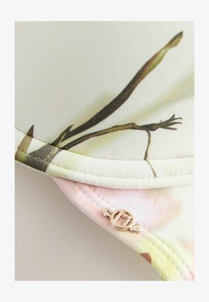 Zbliżenie tkaniny z kwiatowym nadrukiem i małym, okrągłym, metalowym medalionem z logo przyszytym blisko szwu.