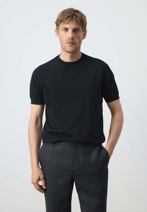 Hombre con un suéter negro de manga corta y cuello redondo y pantalones gris oscuro, de pie con una mano en el bolsillo contra un fondo liso.
