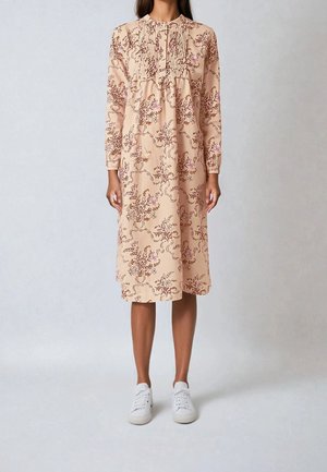 Lang roze jurk met bloemenpatroon en geplooide details bij de halslijn. Draagt witte sneakers, ontwerp tot op de knie.