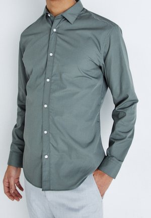 Man draagt een nauwsluitend grijs overhemd met lange mouwen en kraag, gecombineerd met lichtgekleurde broek, staand tegen een effen achtergrond.
