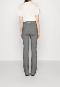 Mujer vestida con pantalones de estampado houndstooth en blanco y negro y una camiseta blanca de manga corta, de pie y mirando hacia atrás frente a un fondo liso.