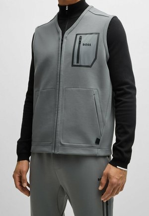 Mann trägt graue Reißverschlussweste mit schwarzem, eingefasstem Reißverschluss-Brusttasche mit der Aufschrift BOSS, über einem schwarzen Langarmshirt und passenden grauen Hosen.