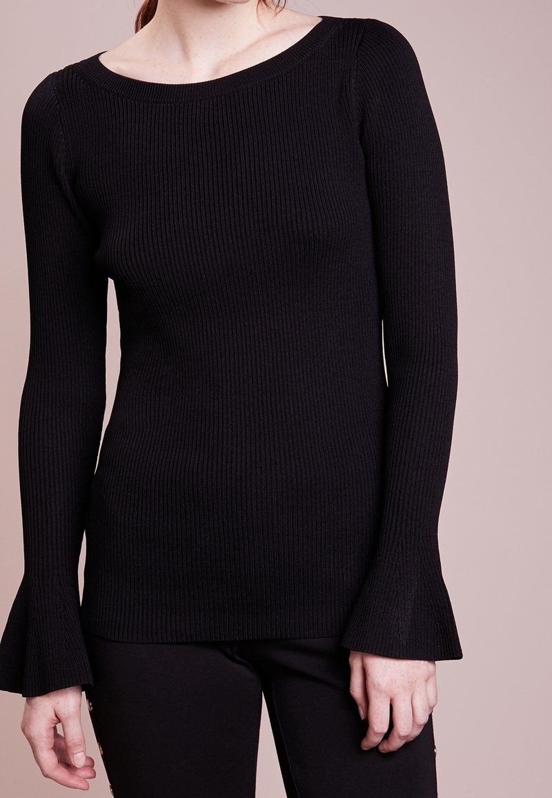 Svart, ribbad stickad långärmad topp med vida, utsvängda manschetter vid handleden och båthals, i kombination med svarta byxor.