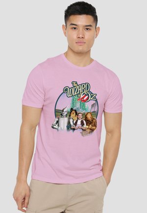 Young man wearing a light pink t-shirt featuring The Wizard of Oz characters and emerald city background.
