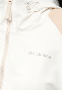 Gros plan d'une veste Columbia couleur écrue avec fermeture éclair, capuche, cordon de serrage et logo sur la zone gauche de la poitrine.