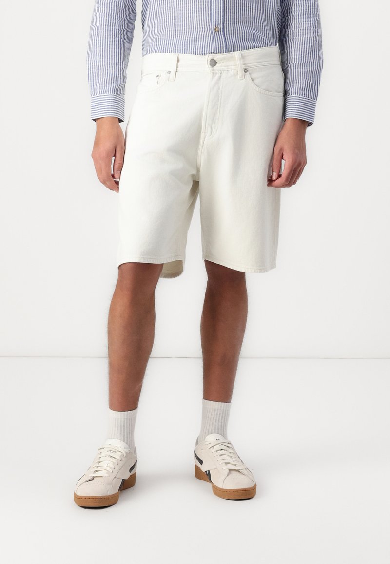 Hvide denimshorts med en afslappet pasform, der har forlommer og knaplukning, parret med lyse sneakers og ankelsokker.