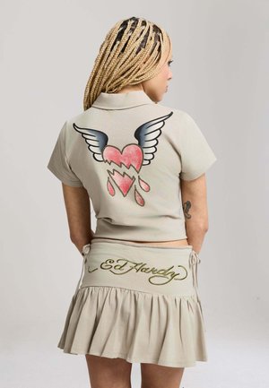 Kvinde med lange blonde fletninger iført beige cropped skjorte med vingeformet, blødende hjerte-grafik og matchende nederdel med "Ed Hardy" broderi.