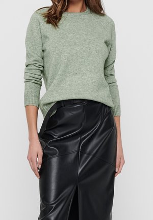 Kvinde iført en lysegrøn strikket sweater, stoppet ned i et sort, højtaljet lædernederdel med en frontsplit og sort bælte.
