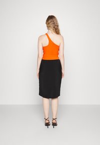 Donna con lunghi capelli mossi, che indossa un top arancione a spalla unica, una gonna nera fino al ginocchio e sandali neri con cinturini, in piedi contro uno sfondo neutro.