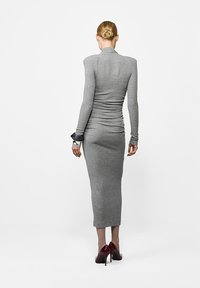 Hall griit, kehaõhuke pikkade varrukatega kleit kõrge kaelusega ja kogutud tekstuuriga. Mudel kannab tumepunaseid kõrge kontsaga kingi, mis rõhutavad sujuvat disaini.