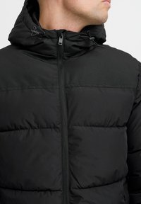 Black puffer jacket with a zip front, hood, and drawstrings. Features a quilted texture and sleek, matte finish material.