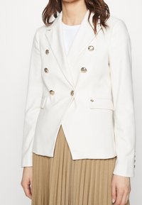 Donna che indossa un blazer bianco doppiopetto con bottoni dorati sopra una maglietta bianca e una gonna plissettata beige, su uno sfondo uniforme.