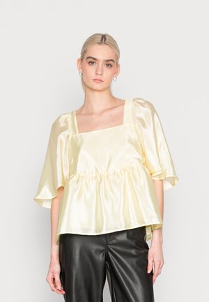 Top amarelo em cetim com decote quadrado, mangas curtas bufantes e cintura franzida. Combinado com calças pretas texturizadas. Design sem emendas.