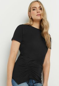 T-shirt nera a maniche corte, con dettagli plissettati sui lati, scollatura rotonda e orlo leggermente curvato. Tessuto morbido e elasticizzato.