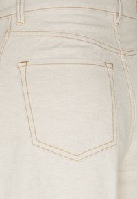 Beige denim fabric featuring a single back pocket with a chevron-shaped design and contrasting yellow stitching. Textured surface visible.
