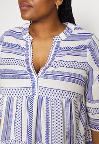Vit topp med blåa geometriska mönster, med en delad halsringning, knappslå och samlad nederkant för en avslappnad passform. Korta ärmar finns.