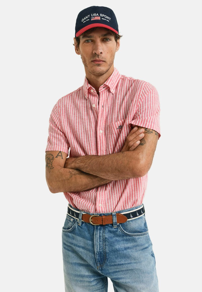 Camicia a maniche corte con bottoni presenta righe verticali rosa e bianche, abbinata a jeans in denim blu e un cappellino blu navy con accenti rossi.