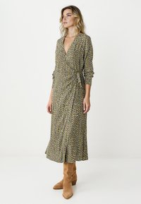 Robe portefeuille fleurie en nuances de jaune, vert et brun. Dotée de manches longues, d'un décolleté en V et d'une silhouette fluide. Portée avec des bottines marron.