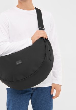 Persona con camisa blanca de manga larga y vaqueros azules sosteniendo un gran bolso negro en forma de media luna con correa ajustable.