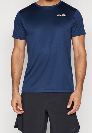 Homme portant un t-shirt bleu marine ellesse et un short noir Nike, debout, les bras détendus le long du corps devant un fond uni.