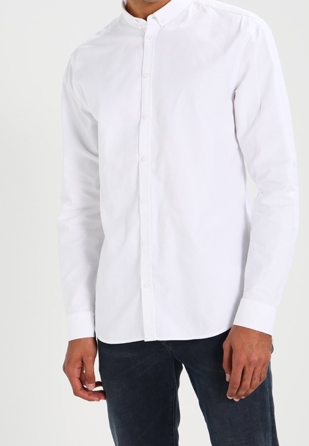 Camicia bianca a maniche lunghe con colletto arrotondato, in morbido cotone, con chiusura frontale a bottoni, indossata sopra jeans di denim scuro.