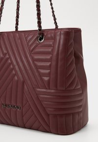 Sac cabas en cuir bordeaux avec un motif matelassé, des poignées en chaîne et un logo en relief. Texture douce avec des détails géométriques à la surface.