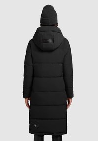 Long black puffer coat with a hood, featuring a quilted texture, side slits, and a branded patch on the back. Paired with a dark beanie.