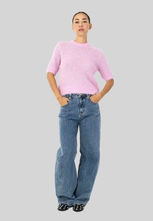 Donna in piedi con le mani in tasca, che indossa un maglione rosa a maniche corte, jeans blu larghi e scarpe nere con borchie su sfondo grigio.