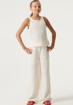 Roomkleurige gebreide tanktop en broek met wijde pijpen, voorzien van een gestructureerde stof. De outfit wordt gecombineerd met felroze teenslippers.