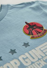 Lys blå bomulds t-shirt med et rødt og sort rundt patch, som har en raket og teksten "WINGMAN", omkranset af mørkegrå stjerner.