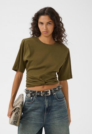 Kvinde med krøllet hår har en olivengrøn cropped t-shirt på, højtaljede blå jeans, sort bælte med sølv øjer, og holder en clutch med slangeprint.