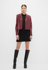 Giacca cropped in ecopelle bordeaux sopra una blouse nera testurizzata, abbinata a una mini gonna nera e stivali alti in pelle nera fino al ginocchio.