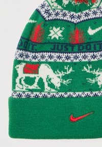 Gorro de malha verde com renas, flocos de neve e árvores em padrão vermelho e branco. Apresenta um swoosh vermelho da Nike perto da parte inferior.