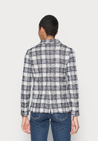 Chaqueta a cuadros en azul marino y blanco, hecha de tejido entrelazado. Presenta un cuello, una abertura en la parte posterior y puños abotonados. Combinada con jeans azul oscuro.